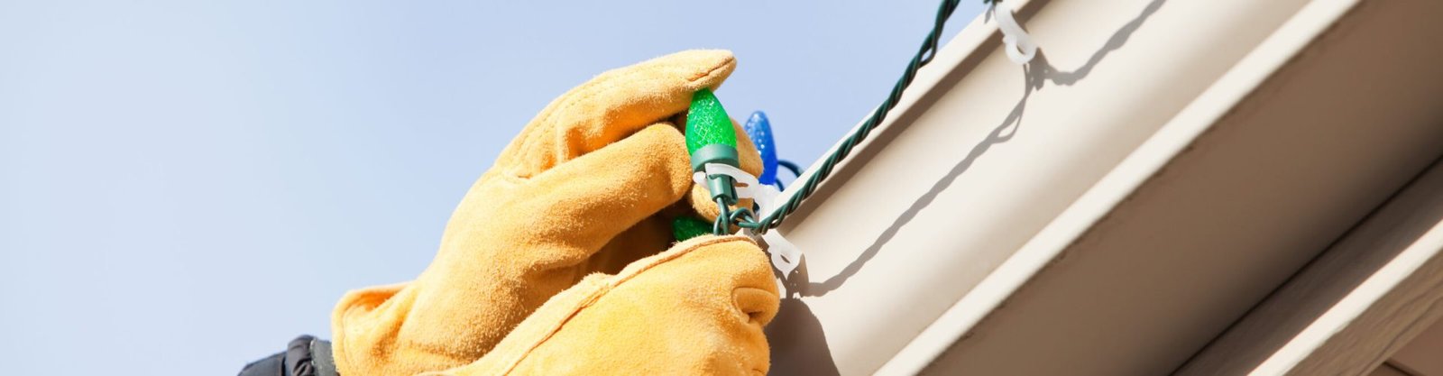 Gloved hands are installing a LED Christmas light onto a clip attached to a house rain gutter. This image could work equally well for a removing concept. The LEDs are housed within faceted plastic covers which replicate the look of incandescent bulbs. LEDs last longer and are much more efficient than conventional bulbs.